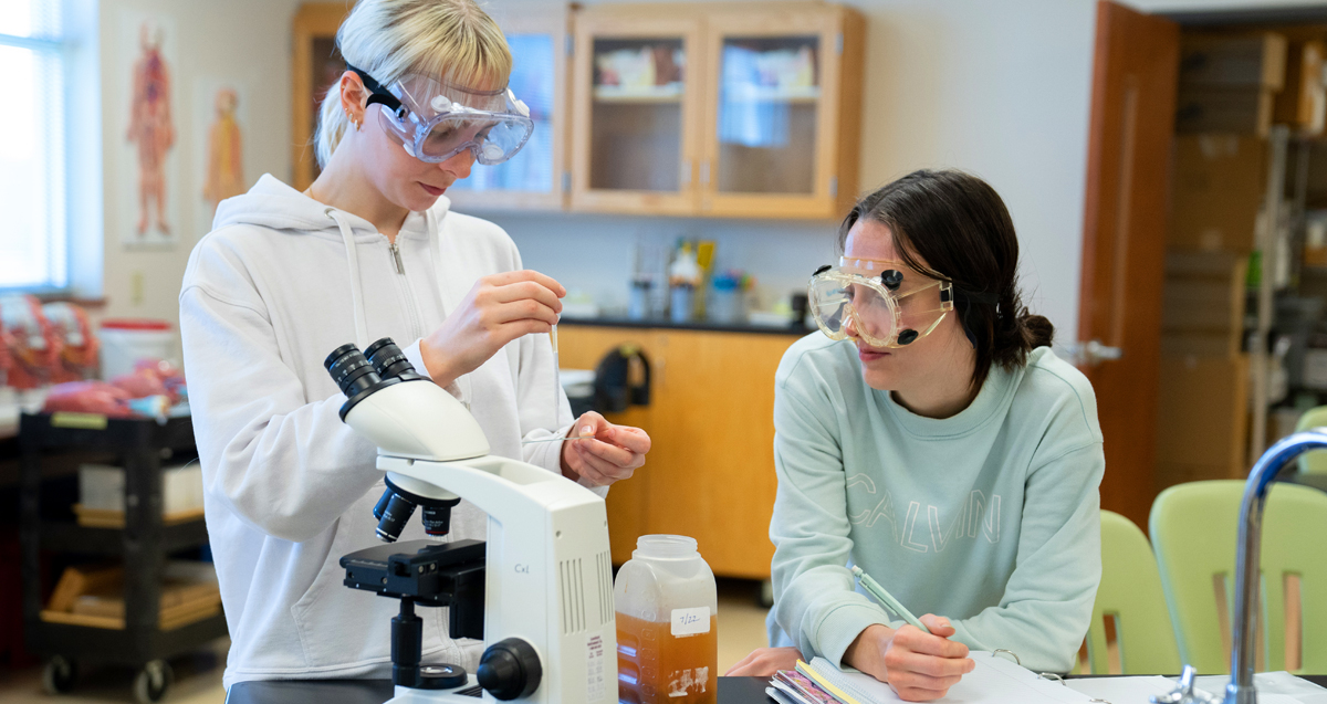 Two students in a laboratory wearing safety goggles. One is using a microscope and the other is taking notes.