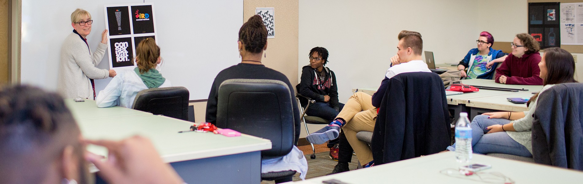 A classroom setting with La Roche University students seated at desks and a teacher standing near a white board, engaging with the students.