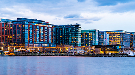 View of modern waterfront buildings at dusk with vibrant lighting and calm water in the foreground.