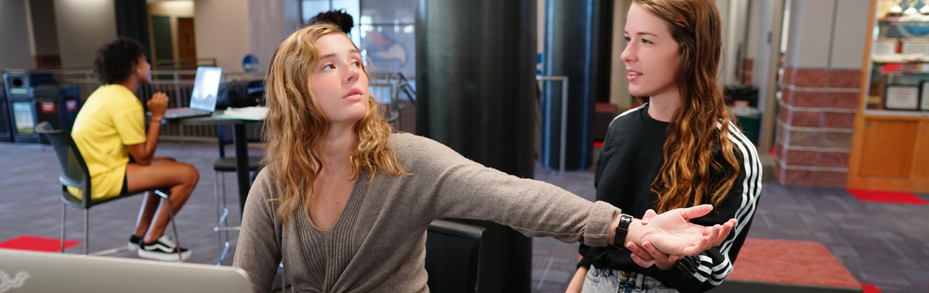 Two La Roche University students practice health assessments, with one sitting at a laptop and gesturing toward the other who is listening attentively. Another person is working on a laptop in the bac