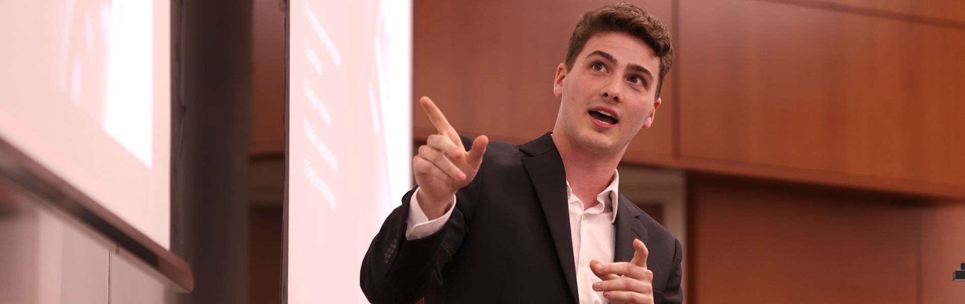 A La Roche University student gives a presentation in a conference room with a projected screen visible in the background. The presenter is speaking and gesturing toward the screen.