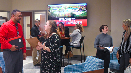 Several individuals engaged in conversation in La Roche University's Admissions Welcome Center, with a welcome sign and a scenic campus photo displayed on digital screens in the background.