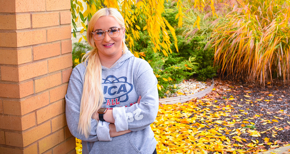 A La Roche University student wearing a hoodie and glasses, smiling, standing next to a brick wall with autumn leaves on the ground.