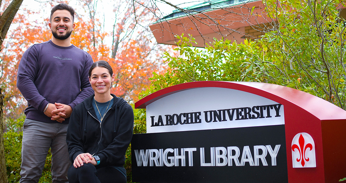 Two individuals smiling in front of La Roche University's Wright Library sign, surrounded by autumn foliage.