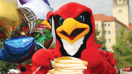 The La Roche University Redhawk mascot sits with a stack of pancakes, surrounded by colorful balloons with a campus building in the background.