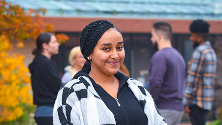 A young woman is standing outdoors, smiling at the camera. Behind her, several people are casually talking in small groups
