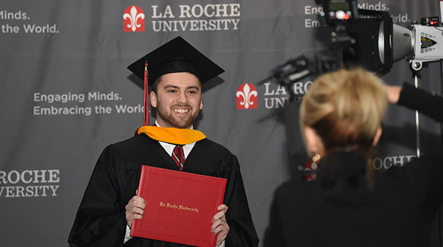 A professional photographer take a photo of a La Roche University graduate holding his diploma. The graduate is wearing his cap and gown, and a La Roche University backdrop is behind him.