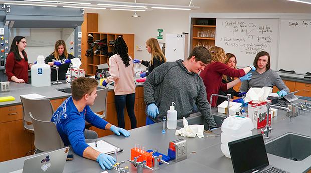 A group of students in a biochemistry class work in the science laboratory at La Roche University. Some students wear blue gloves. One student is placing items in a sink.
