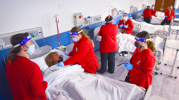 Nursing students wearing face shields and red jackets practice check vitals on a mannequin in the simulation lab at La Roche University. 