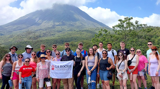 A group of La Roche University students standing in front of the Arenal Volcano in a lush landscape.