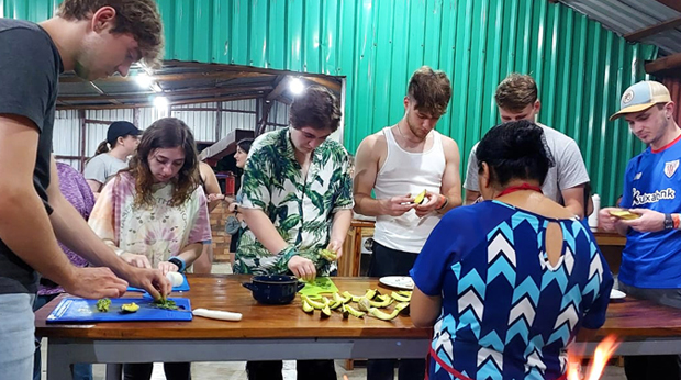 Group of people engaged in a cooking class, preparing food together at a large table with various ingredients and a stove visible.