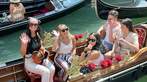 Five La Roche students enjoy a sunny day on a gondola ride in Venice, waving and smiling as they pass.