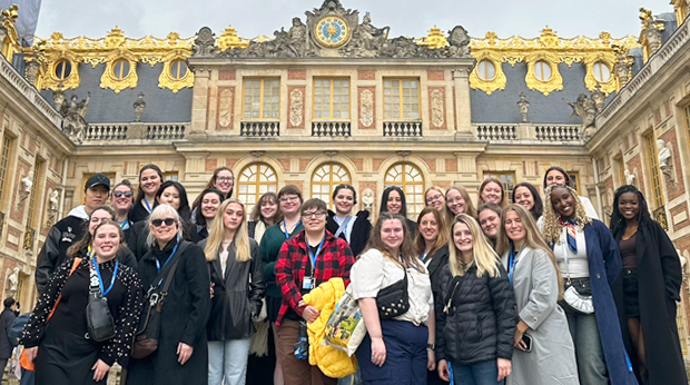 A group of La Roche University students posing in front of the Palace of Versailles, with its ornate golden architectural details visible in the background.