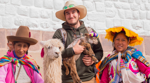 A La Roche University student holding a baby alpaca stands between two individuals wearing traditional Peruvian attire including vibrant textiles and hats, with a stone wall in the background.