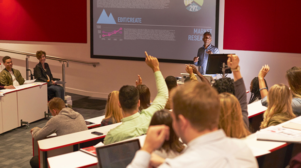 Students in an auditorium classroom. Some are raising their hands. An instructor stands in the center of the room near a projection screen.
