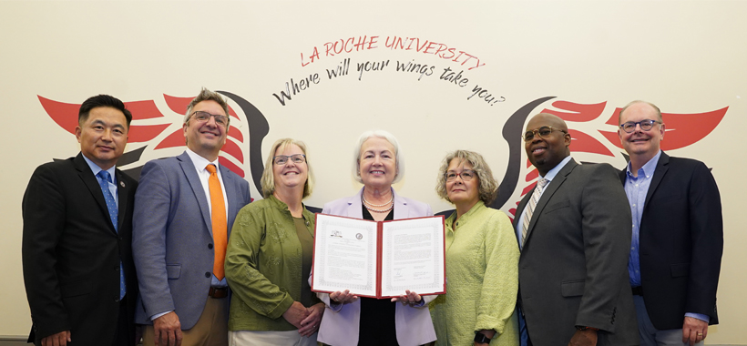 Group of seven individuals standing side by side with the La Roche University “Where will your wings take you?” wings graphic in the background. One person is displaying a certificate towards the camera. They are dressed in business attire. Group of seven individuals standing side by side with the La Roche University “Where will your wings take you?” wings graphic in the background. One person is displaying a certificate towards the camera. They are dressed in business attire.