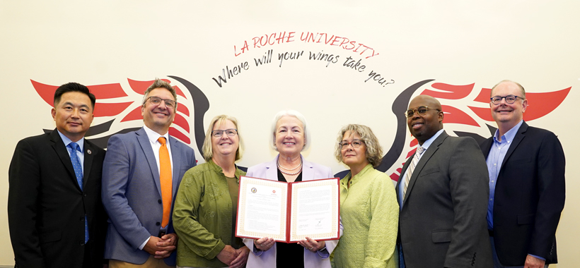 Group of seven individuals standing side by side with the La Roche University “Where will your wings take you?” wings graphic in the background. One person is displaying a certificate towards the camera. They are dressed in business attire. Group of seven individuals standing side by side with the La Roche University “Where will your wings take you?” wings graphic in the background. One person is displaying a certificate towards the camera. They are dressed in business attire.