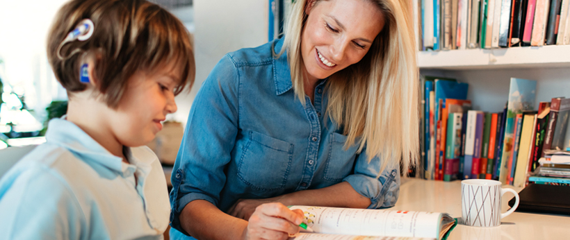 Teacher assisting a studentat at a table wearing a hearing device with his workbook exercise.