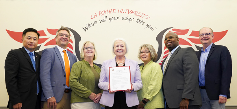 Group of seven individuals standing side by side with the La Roche University “Where will your wings take you?” wings graphic in the background. One person is displaying a certificate towards the camera. They are dressed in business attire. Group of seven individuals standing side by side with the La Roche University “Where will your wings take you?” wings graphic in the background. One person is displaying a certificate towards the camera. They are dressed in business attire.