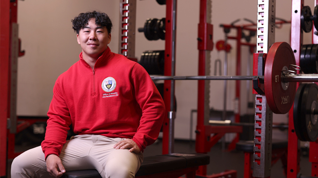 La Roche University alumnus sits on a bench in the weight room of the Kerr Fitness & Sports Center with weight racks and barbells in the background.