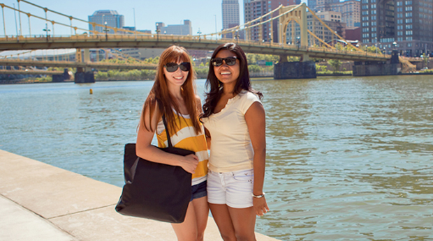 Two La Roche University students on the North Shore of downtown Pittsburgh in the summertime. The river and a yellow bridge are behind them.