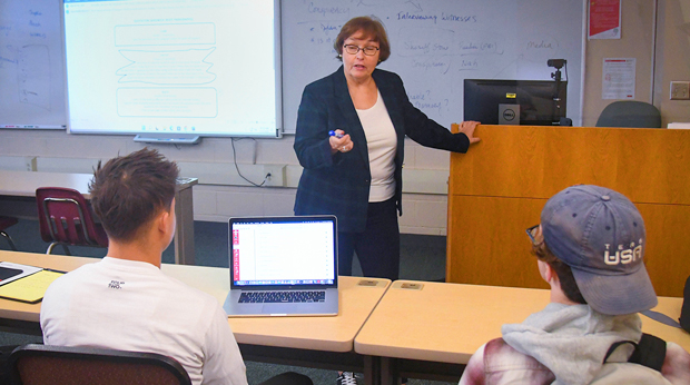 A professor at La Roche University lectures in front of a large screen and white board. Two students sit in front row of the classroom. One has a laptop open in front of him.