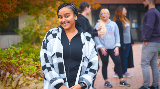 A student wearing a black and white jacket stands on the walkway in front of La Roche University's Wright Library. Four students talk to each other behind her.