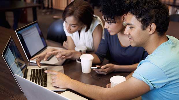 Three students sit at a desk with laptops and disposable coffee cups.