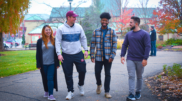 Four La Roche University students laugh together while walking outside on an autumn day. The Zappala Campus Center building and are trees are behind them.