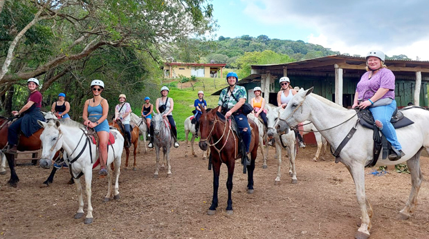 A group of La Roche students on horseback in a rural setting, wearing helmets and riding gear, ready for a trail ride.