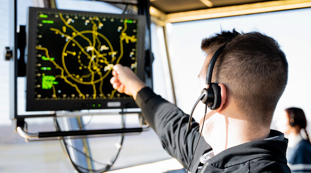 A male student pointing at the air traffic control radar screen. He is wearing a headset and sitting in an air traffic control tower. There is a female student wearing a headset in the background.`