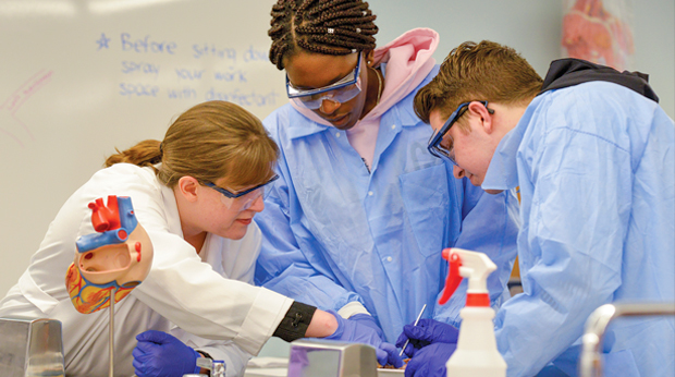 A La Roche University professor assists two students in the anatomy lab. All three are wearing protective eyewear, gloves and blue or white lab jackets.