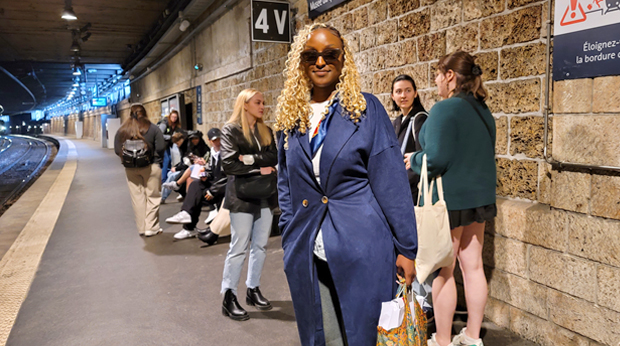 Person wearing sunglasses and a navy blue blazer stands smiling on a platform at a train station, with other passengers waiting in the background.
