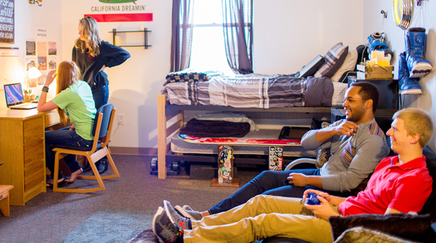 A group of resident students hang out in a room of Bold Hall at La Roche University. Two of them are at a desk looking at a laptop together. The other two students sit on couch playing video games.