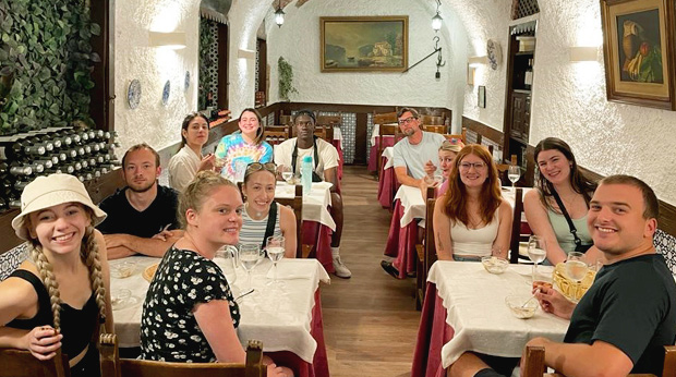 Group of La Roche students enjoying a meal at a traditional restaurant in Spain, sitting around tables arranged in a cozy stone-walled room decorated with paintings.
