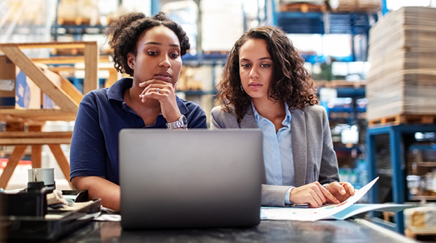 Two workers in a warehouse look at a laptop together.