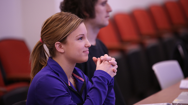 A student sits in a classroom at La Roche University. Her hands are folded and she looks straight ahead. A classmate is next to her in the background.
