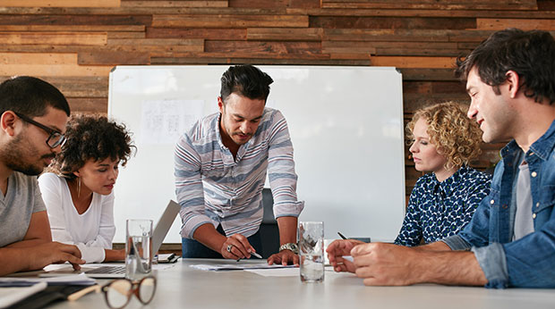 A group of people look at documents together at a conference table. One person is standing and pointing to the documents. The others are sitting.