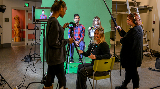 Two La Roche University students use a camera to record a professor in front of a green screen. Another professor sits in a chair next to the camera tripod.