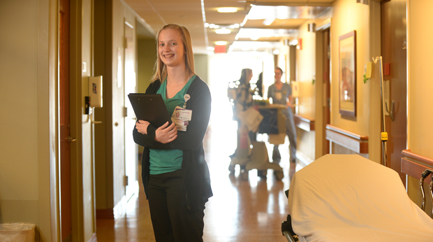A nurse stands in hospital hallway holding a clipboard.