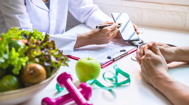 A nutritionist gives a health consultation to patient. The nutritionist points to a smartphone with a pen. They sit at a table with fruits, vegetables, weights and measuring tape.