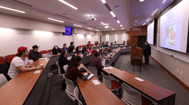 La Roche University students sit at desks in an auditorium lecture classroom. A professor stands in front of a Smart Board.