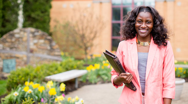 A student carrying a binder stands in front of Magdalen Chapel at La Roche University. There are flowers next to her and in the background.