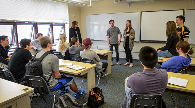 Four students deliver a presentation in a classroom at La Roche University. The rest of the class is seated watching them, and a professor is standing to the side of the room.