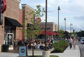 title="Groups of people dining outside at tables with red umbrellas at Hello Bistro. Two people walk on a sidewalk near the restaurant."