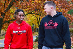 Two La Roche University students, one in a red Redhawks hoodie and the other in a black hoodie with a unique logo, smiling at each other in a park with colorful autumn leaves in the background.