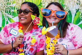 Two La Roche University students wearing colorful tie-dye shirts while holding tropical themed paper props and drinks at a festive event. One person sports oversized orange sunglasses.
