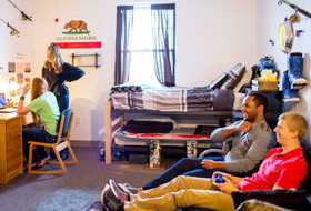 Four La Roche University students in a residence hall room. Two are studying at a desk and the other two are playing video games.