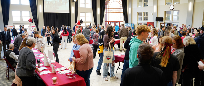 A crowd of prospective students and parents at an Open House event at La Roche University. There are tables covered in red tablecloths with various brochures and fliers laid out on top. There are professors and students conversing in the background.