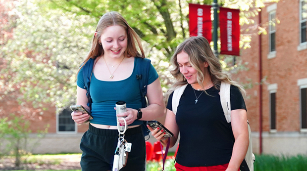 Two individuals dressed in summer attire walking and conversing while looking down at their phones. The individual on the left is holding a phone, keys and a canned beverage. The individual on the right is holding a phone and is wearing a backpack. There are flowering trees, campus banners and a brick building in the background.
